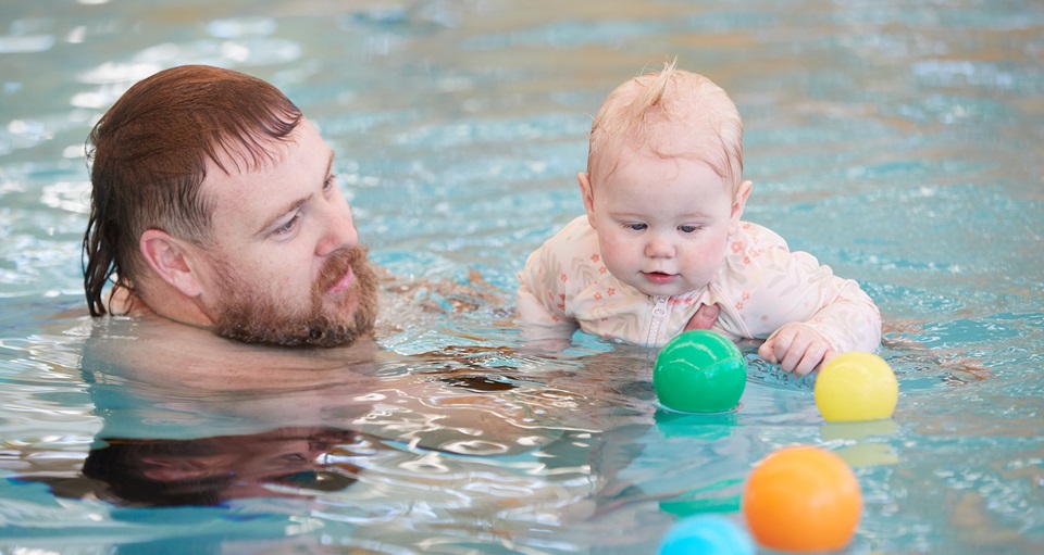 Father with his baby daughter playing with coloured toy balls in a parent baby swim class